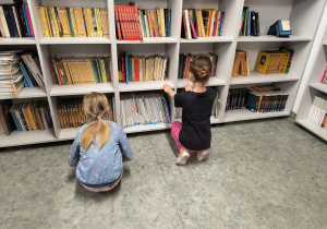 Dzieci kucają przy regale i oglądają książki w bibliotece szkolnej.