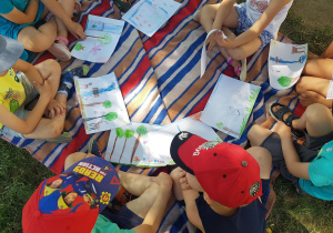 Dzieci siedzą na kocu w ogrodzie, oglądają prace plastyczne.