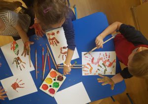 Dzieci siedzą przy stole, na którym są rozłożone farby. Dzieci malują dłonie farbami i tworzą na kartonach barwne kompozycje.