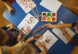 Dzieci siedzą przy stole. Malują dłonie farbami. Na stoliku widać kolorowe prace.