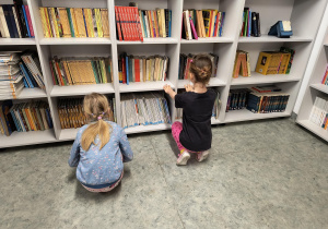 Dzieci kucają przy regale i oglądają książki w bibliotece szkolnej.