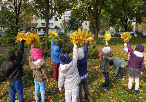 Grupa dzieci stoi w ogrodzie. Dzieci trzymają zrobione przez siebie bukiety z liści.