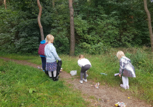 Grupa czterech dziewczynek w parku. Jedna stoi na ścieżce, pozostałe zbierają porozrzucane papiery.