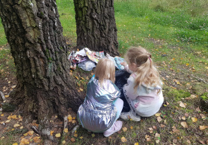Dwie dziewczynki kucają przy drzewie i podnoszą śmieci z trawnika.