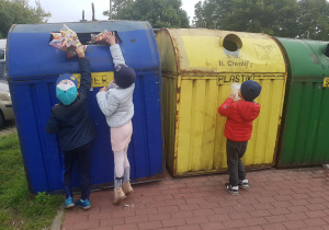 Dzieci stoją przy pojemnikach do segregowania odpadów. Dwóch chłopców wrzuca gazety do pojemnika niebieskiego. Jeden chłopiec wrzuca plastikowe opakowanie do pojemnika żółtego.