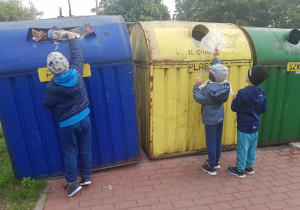 Dzieci stoją przy pojemnikach do segregowania odpadów. Chłopiec z lewej strony wrzuca do niebieskiego pojemnika papier. Dwóch chłopców z prawej strony wrzuca do żółtego pojemnka folię.