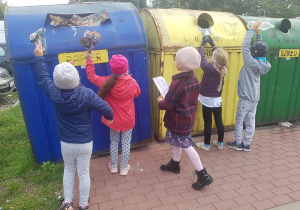 Dzieci stoją przy pojemnikach do segregowania odpadów. Wrzucają do nich znalezione śmieci.