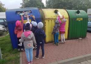 Dzieci stoją przy pojemnikach do segregowania odpadów. Dziewczynki i chłopcy wrzucają do pojemników plastikowe torebki, gazety, papierowe opakowania.