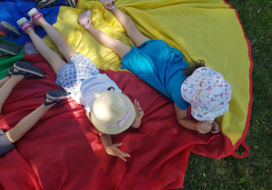 Dzieci w trakcie zabaw relaksacyjnych leżą na kolorowej chuście animacyjnej w ogrodzie przedszkolnym.