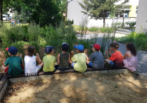 Dzieci siedzą na obrzeżu piaskownicy i uczestniczą w ćwiczeniach relaksacyjnych.