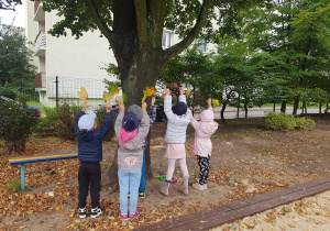 Grupa dzieci stoi przy pniu dużego drzewa. Mają uniesione do góry ręce, trzymają w nich liście.