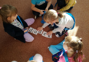 Dzieci siedzą na dywanie i układają napis dydek.