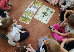 Dzieci siedzą na dywanie i oglądają ilustracje przedstawiające drzewa.
