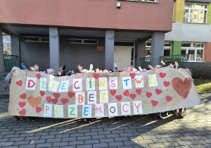 Dzieci stoją przed przedszkolem i prezentują zbiorową pracę plastyczną.